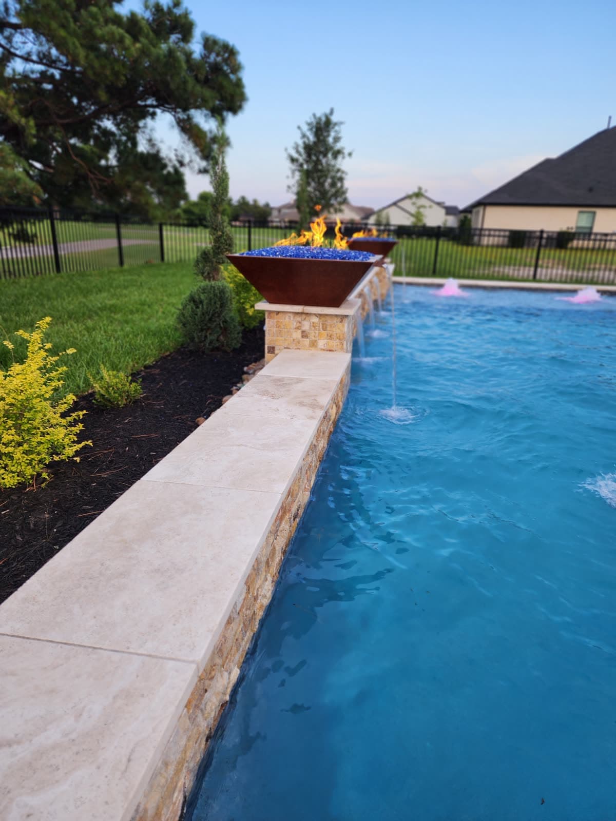 Custom pool in Cypress, TX. At dusk, two copper fire bowls burn over blue fire glass while water spills beneath them and the pool lights turn the surface warm.