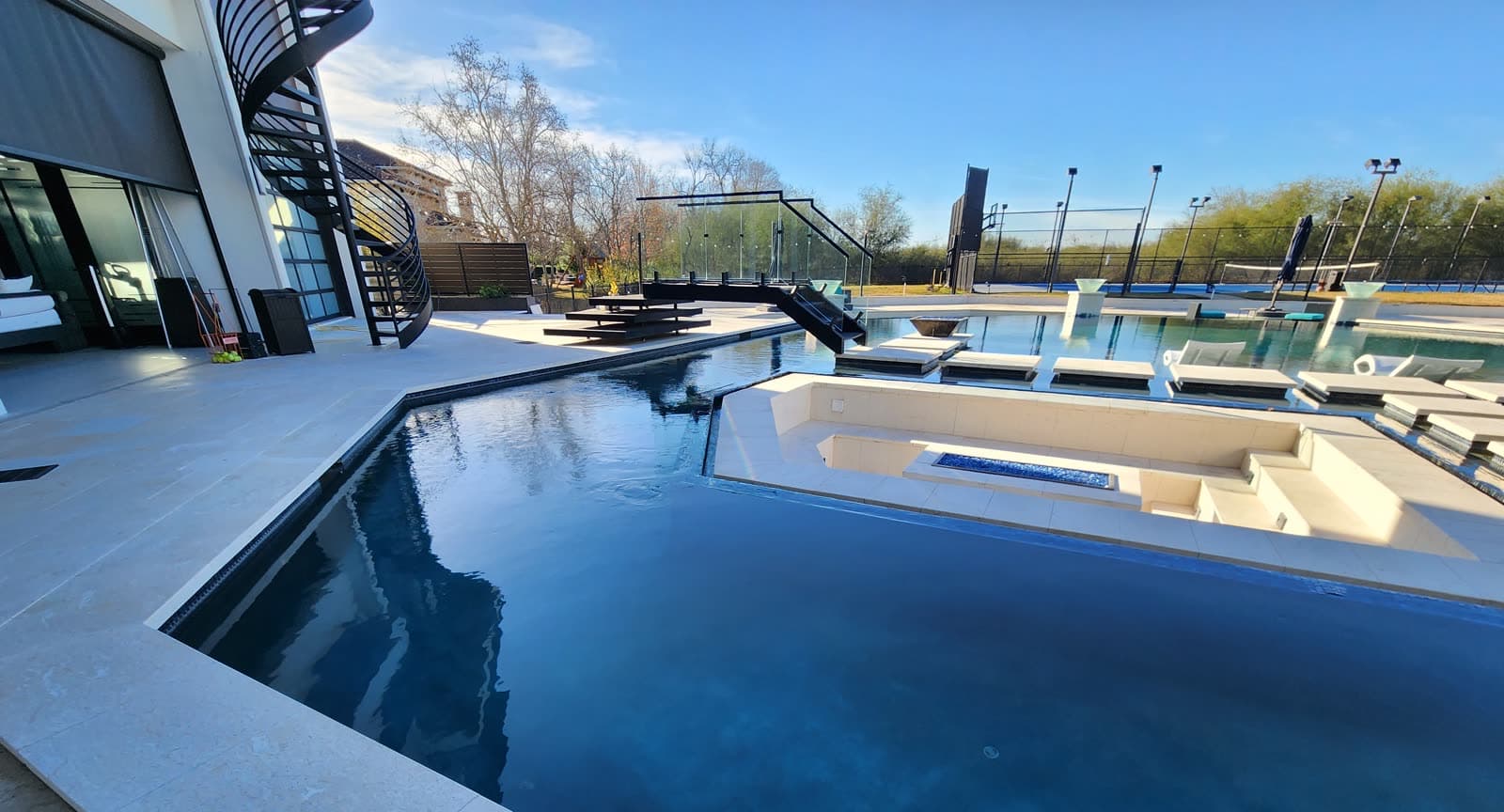 Custom pool in Richmond, TX. A glass-decked bridge with glass railings crosses a dark-water pool, in-water loungers and a sunken fire feature beside it.