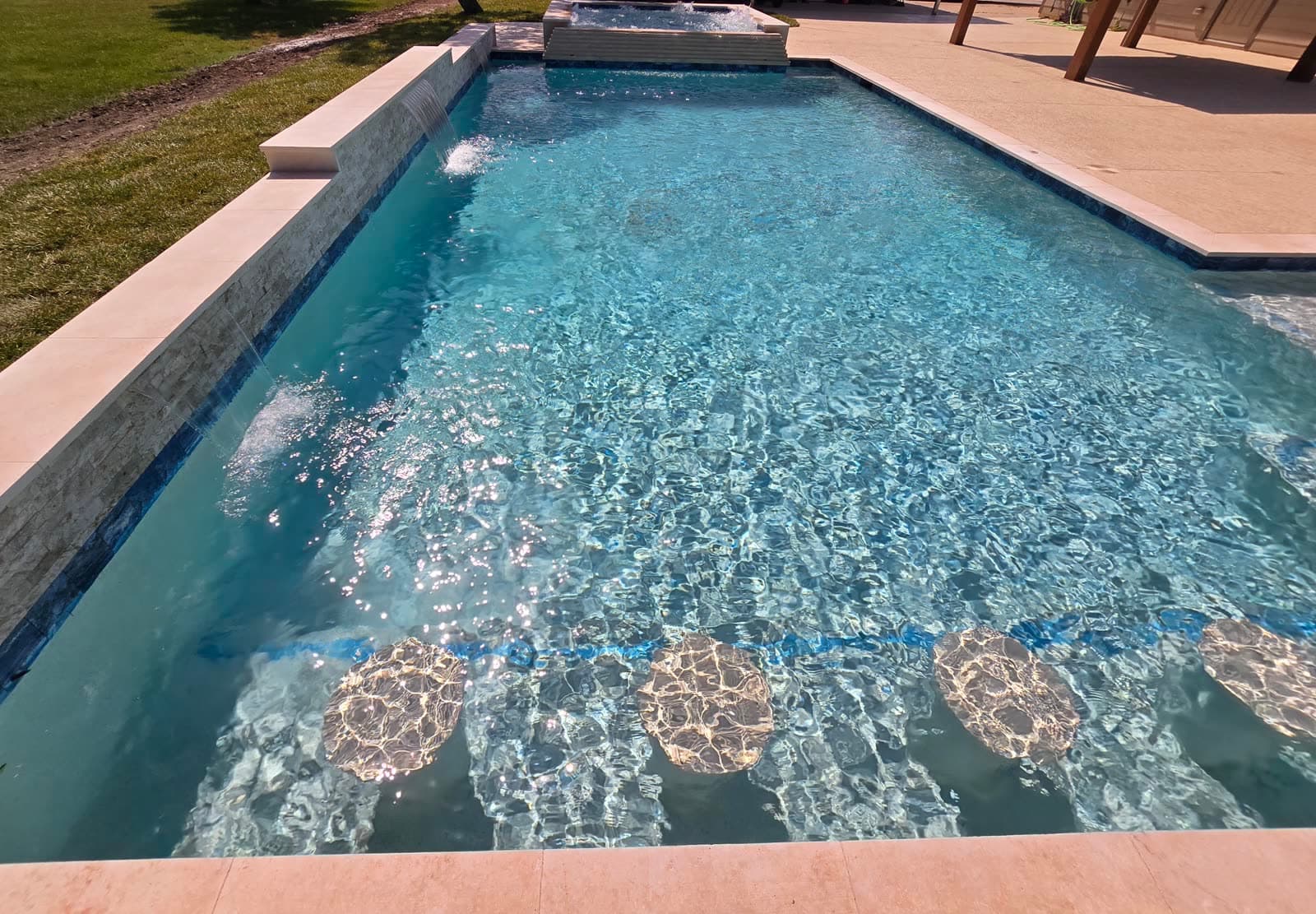 Custom pool in Richmond, TX. A volleyball pool in full sun: a line of blue tile with round medallions crosses the floor, water sheets down the left wall, and the built-in spa bubbles at the far end.