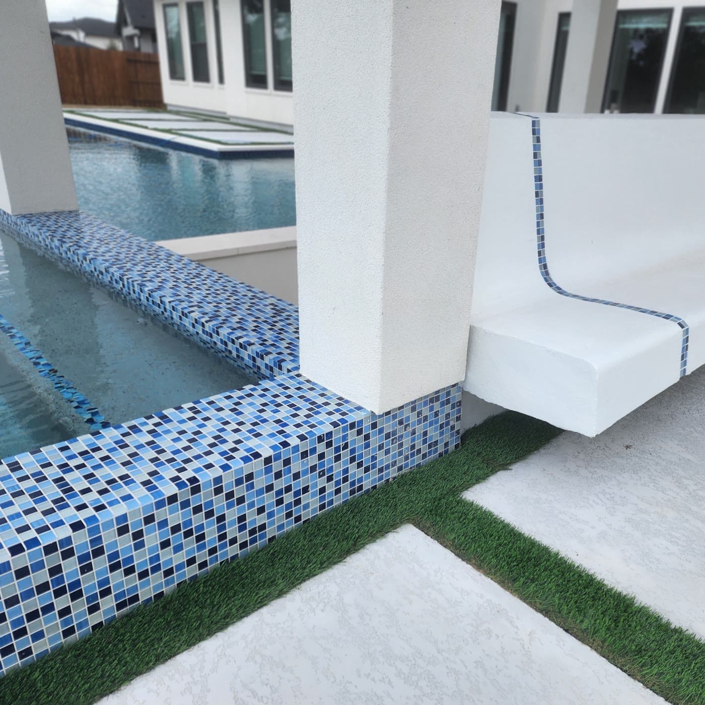 Inground pool in Cypress, TX. A raised pool edge clad in three tones of blue glass mosaic, with a cantilevered bench beside it and turf strips set between the pavers.